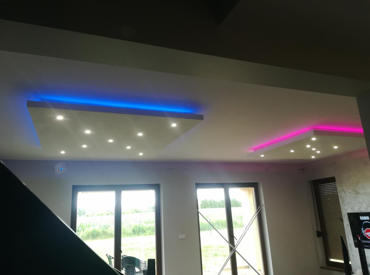 Nowoczesny salon z podwieszanym sufitem, oświetleniem punktowym i dekoracyjnym podświetleniem LED w kolorach niebieskim i różowym, widok na okna z zielenią na zewnątrz.