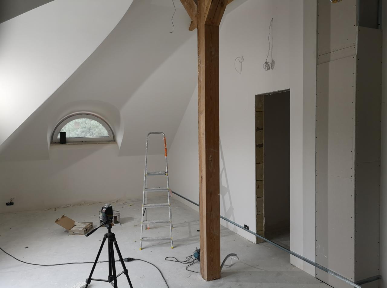 Poddasze w trakcie wykańczania: widoczna drewniana belka konstrukcyjna, okrągłe okno, drabina aluminiowa, laser krzyżowy na statywie, częściowo zabudowana ściana z płyt kartonowo-gipsowych...