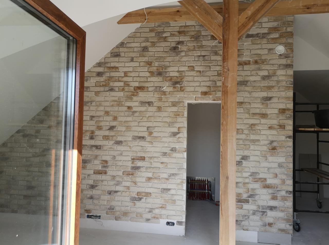 Ściana wyłożona dekoracyjną cegłą w odcieniach beżu i brązu, z widoczną drewnianą belką konstrukcyjną i fragmentem okna, na poddaszu ze skosami.