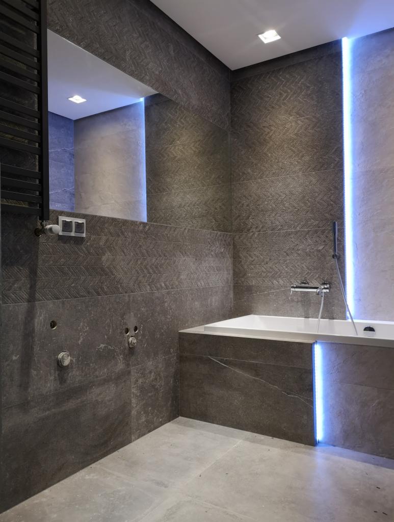 Nowoczesna łazienka w odcieniach szarości z podświetlaną wanną i dużym lustrem, widoczne niedokończone podłączenia hydrauliczne.