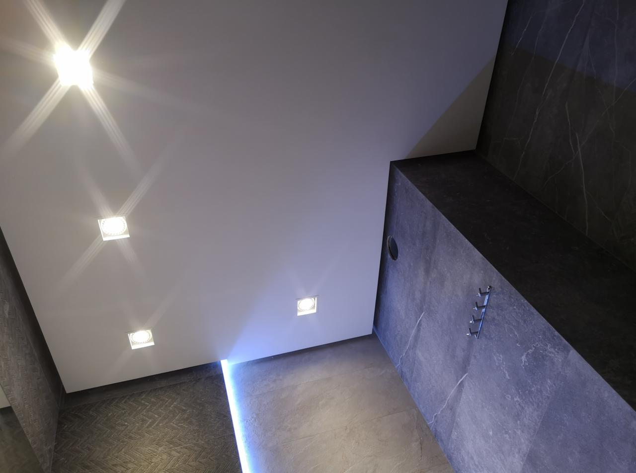 Nowoczesna łazienka z białym sufitem podwieszanym, oświetleniem punktowym i dekoracyjną listwą LED, ściany wykończone płytkami imitującymi marmur, widok z dołu.