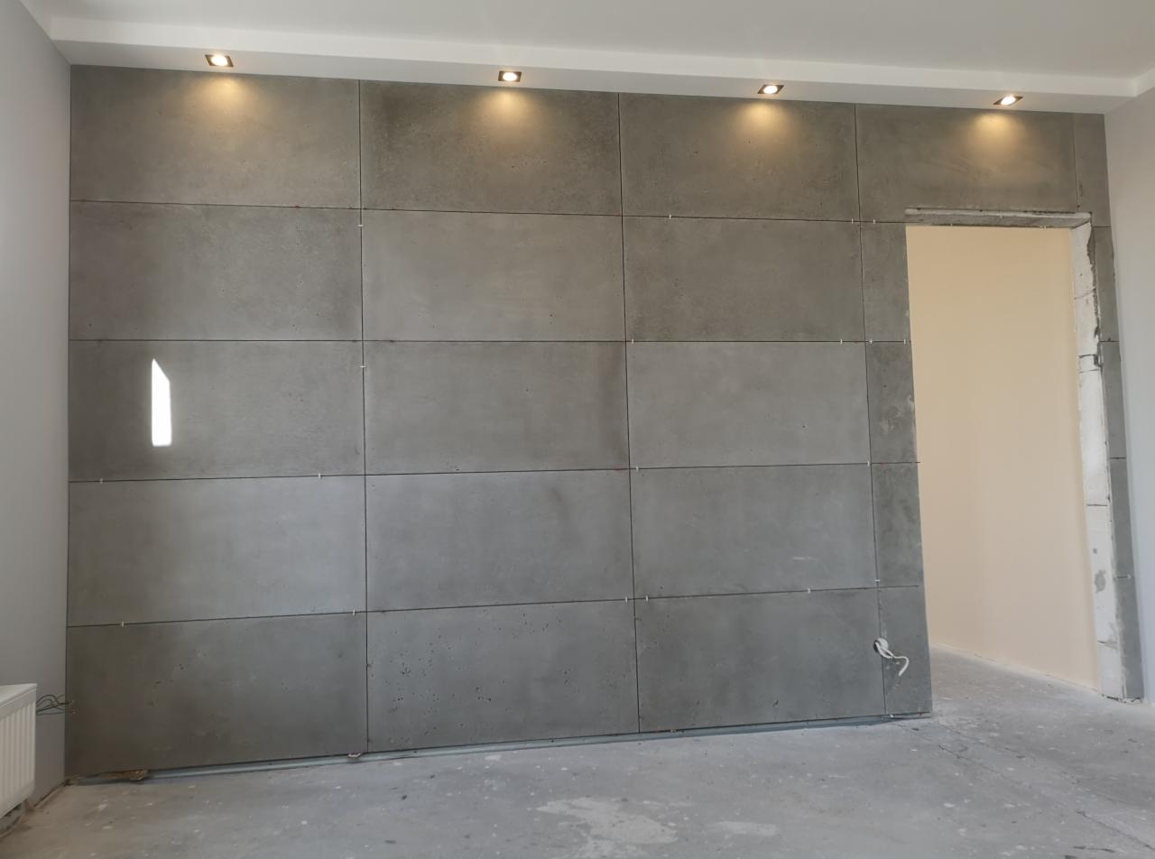 Ściana wyłożona dużymi, szarymi płytami imitującymi beton, oświetlona punktowymi lampami sufitowymi, z widocznym otworem drzwiowym w stanie surowym.