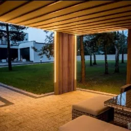 Nowoczesna, oświetlona altana z drewnianą konstrukcją i meblami ogrodowymi, widok na zadbany trawnik i dom w tle, oświetlenie LED podkreśla pionowe elementy drewniane.