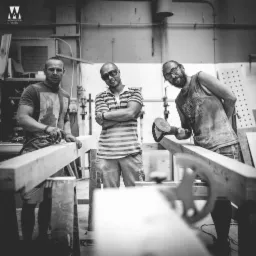 Trzech mężczyzn w warsztacie obrabia drewniane belki szlifierkami, zdjęcie czarno-białe, z widocznym logo studia na ścianie.