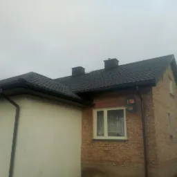 Dom w Raciążu z nowym, ciemnym dachem z blachy, widoczne kominy, rynny i elewacja z cegły oraz fragment ocieplonej ściany.