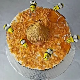 Okrągły tort z dekoracją imitującą ul i pszczoły, posypany płatkami kukurydzianymi, umieszczony na srebrnej paterze.