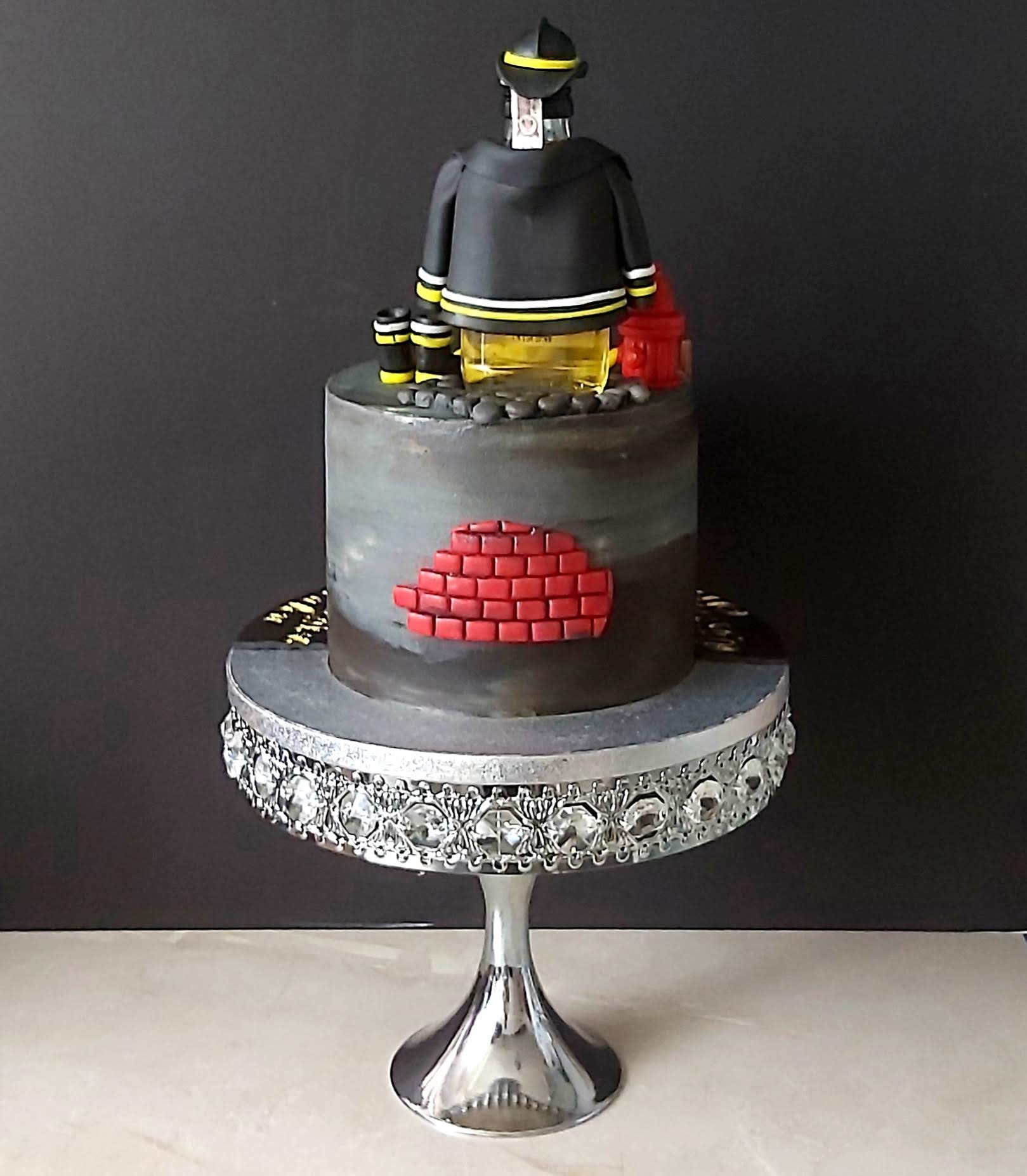 Tort w stylu strażackim, z figurką strażaka, hydrantem i fragmentem muru z czerwonej cegły, na srebrnej paterze.