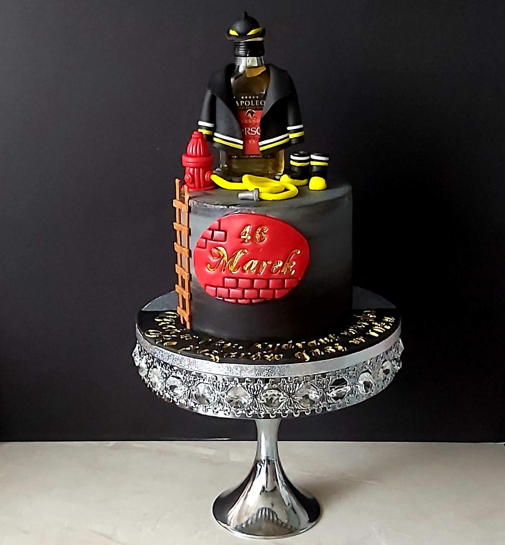 Ozdobny tort na urodziny z motywem strażaka: butelka alkoholu w ubraniu strażackim, hydrant, drabina i napis '46 Marek' na tle imitującym mur z cegieł, całość na srebrnym stojaku.