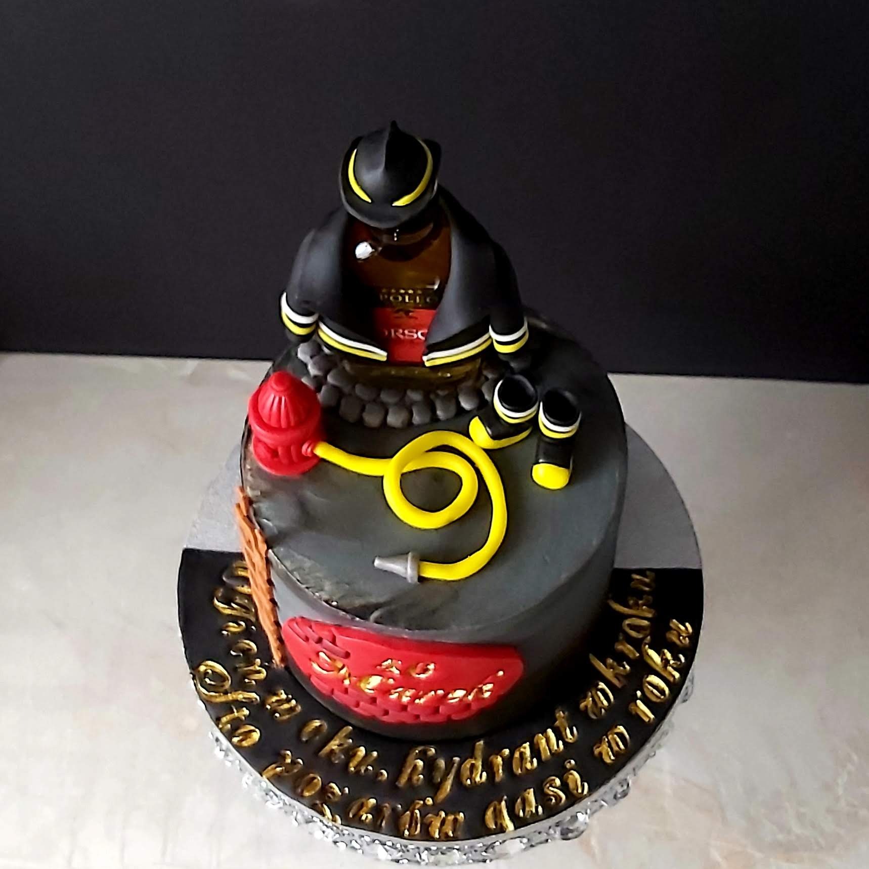 Tort urodzinowy w kształcie strażaka z hełmem, wężem strażackim i hydrantem, butelka alkoholu w roli korpusu, na czarnym podeście z napisem wykonanym złotą czcionką.