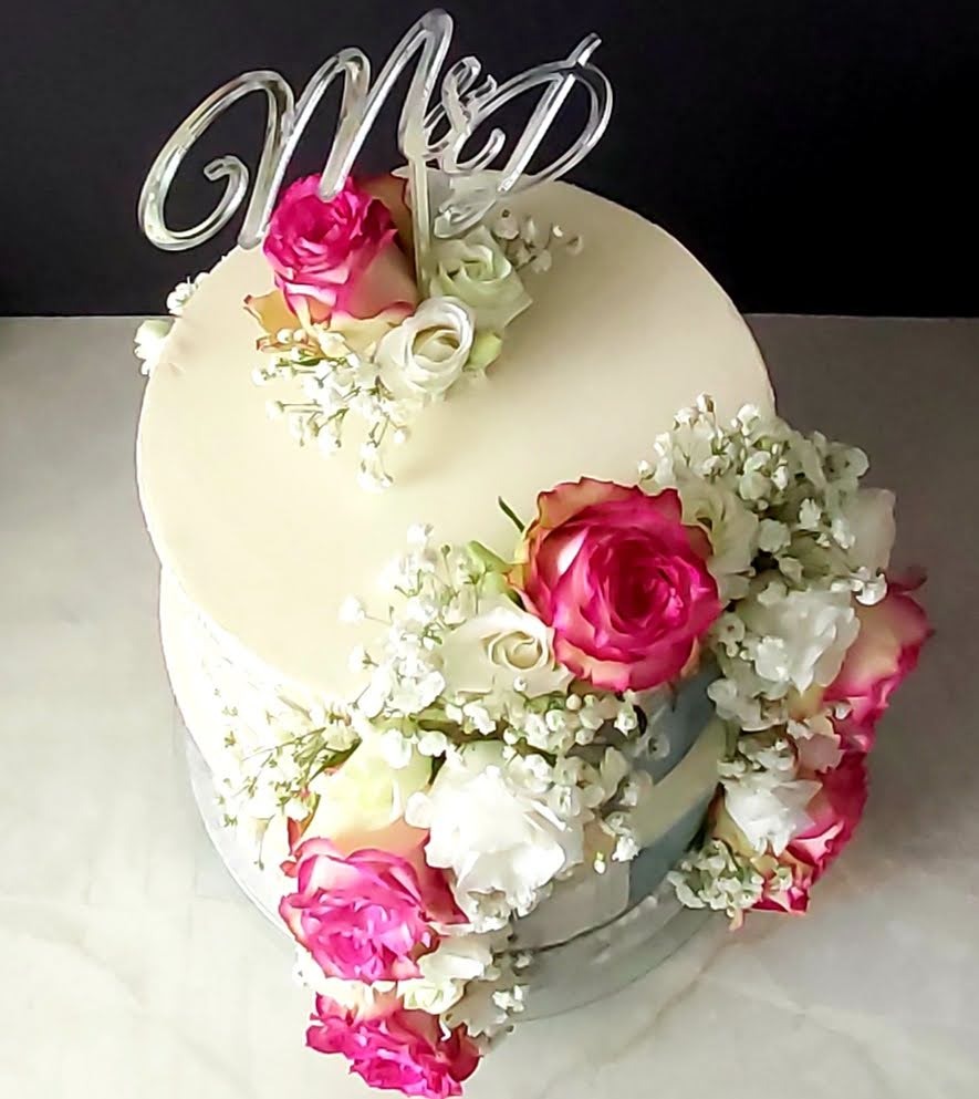 Tort dwuwarstwowy z kremową polewą, dekorowany różowymi i białymi różami oraz gipsówką, zwieńczony akrylowym topperem z inicjałami 'M&D'.