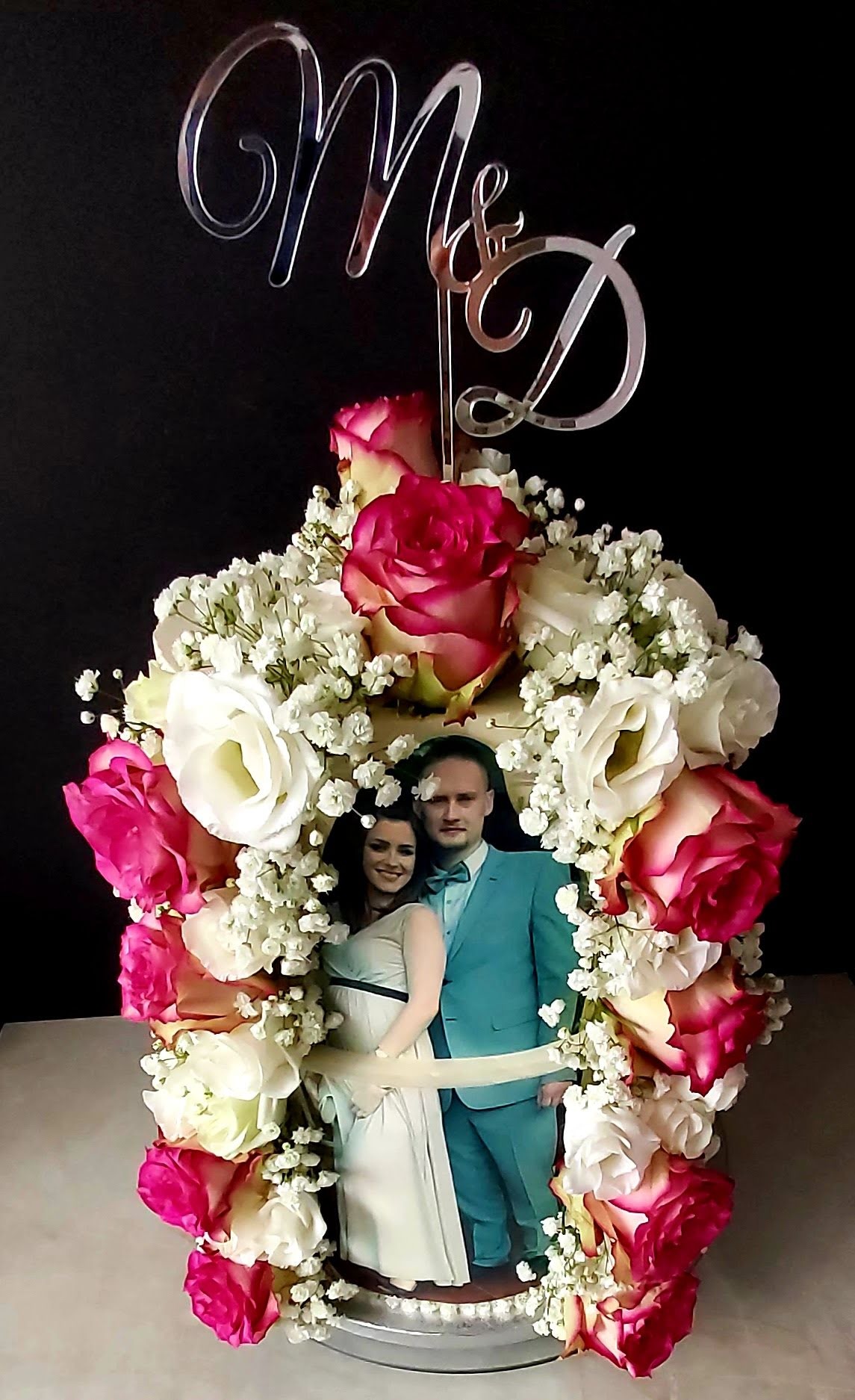 Tort weselny z fotografią pary młodej, otoczony białymi i różowymi różami oraz gipsówką, zwieńczony srebrnym napisem 'M & D'.