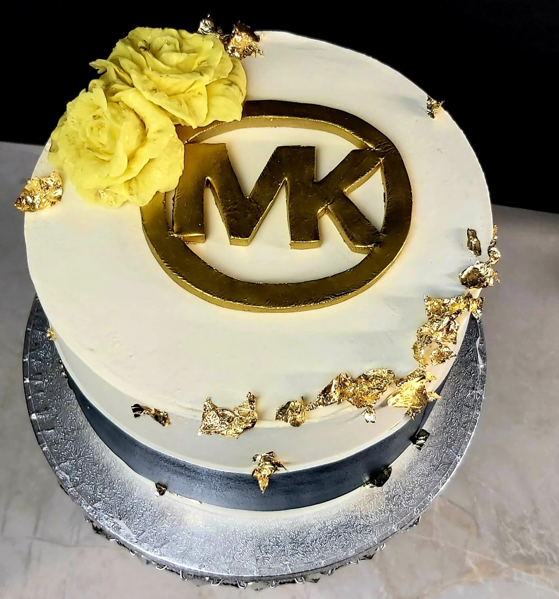 Tort z białym kremem, złotym logo MK, jadalnymi płatkami złota i żółtymi kwiatami z kremu, umieszczony na srebrnej paterze.