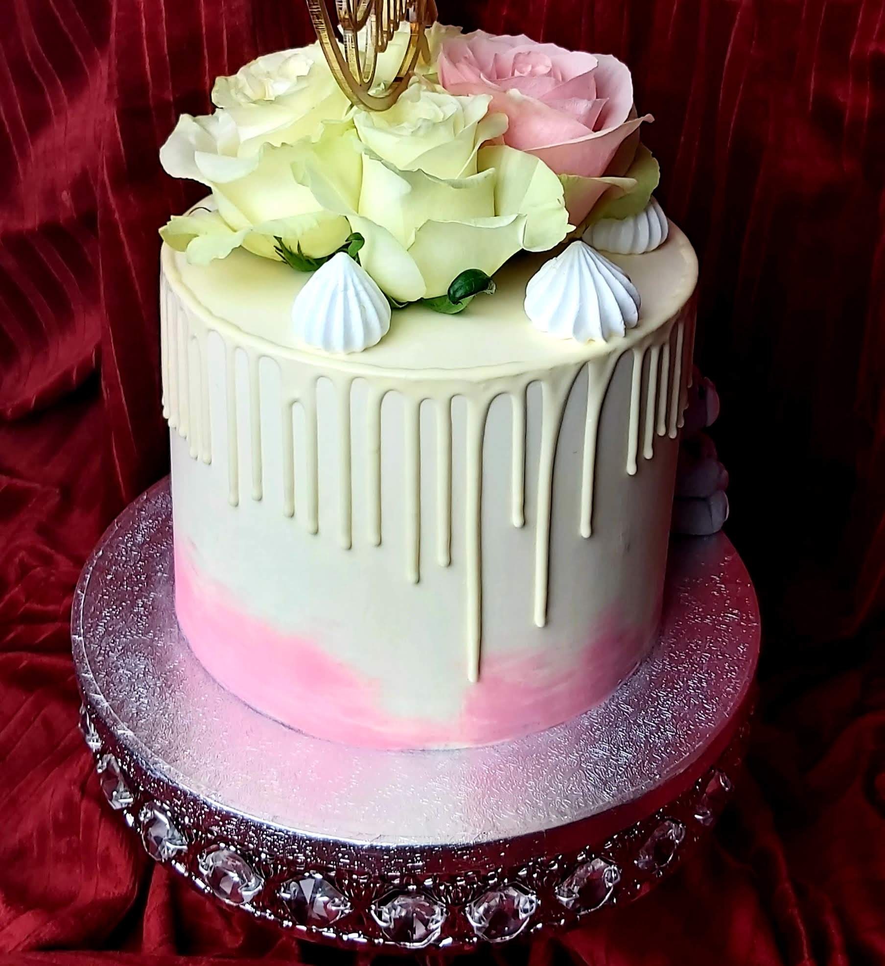 Tort z białym lukrem spływającym po bokach, udekorowany białymi i różowymi różami oraz małymi bezami, na srebrnej paterze z kryształkami, na tle czerwonej, aksamitnej tkaniny.