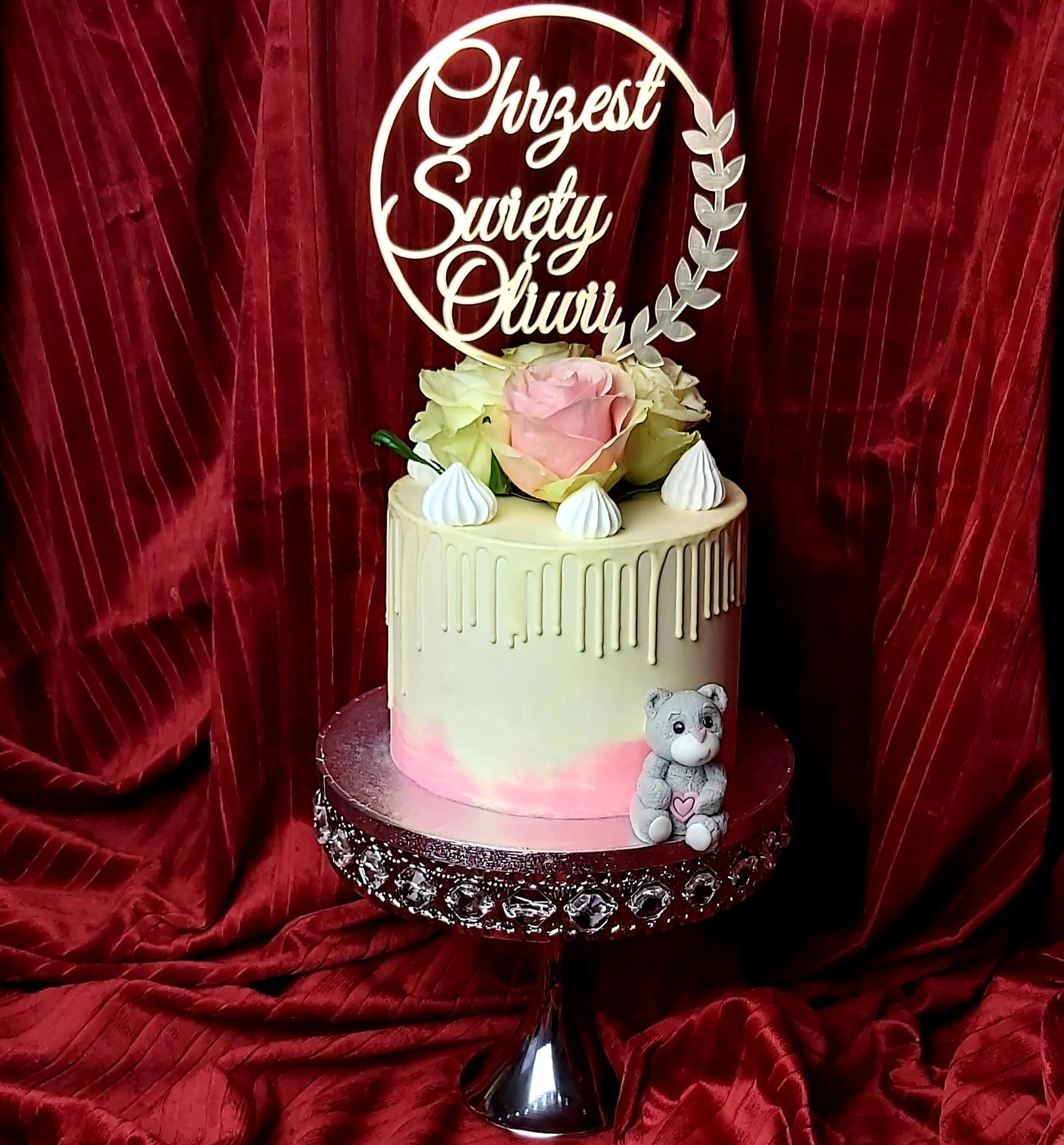 Tort na Chrzest Święty z topperem, różami, bezami i figurką misia na srebrnej paterze z kryształkami, na tle bordowej draperii.