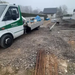 Teren budowy w trakcie przygotowania fundamentów: żwir, zbrojenia, częściowo zalany fundament, w tle budynek i auto dostawcze z logo firmy.