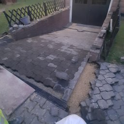 HARAZIN Michał Harazin - Układanie kostki brukowej na podjeździe do garażu. Widoczny fragment pracownika w odblaskowej kamizelce i worki z materiałem. Częściowo ułożona kostka i zamontowany drenaż.