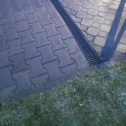 HARAZIN Michał Harazin - Szara kostka brukowa ułożona w regularny wzór, z czarnym odpływem liniowym i fragmentem trawy w rogu. Widoczny cień osoby.