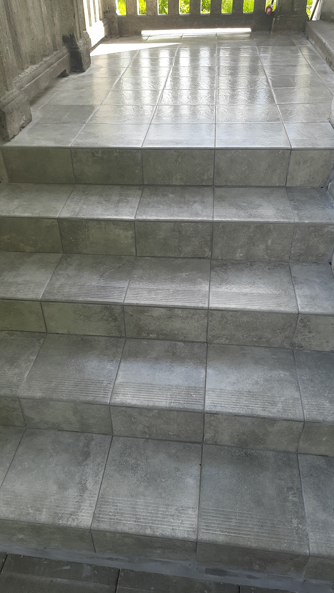 Schody wyłożone szarą, kwadratową kostką brukową z delikatnym wzorem, prowadzące na podest. Widoczne elementy balustrady i refleksy światła na powierzchni kostki.