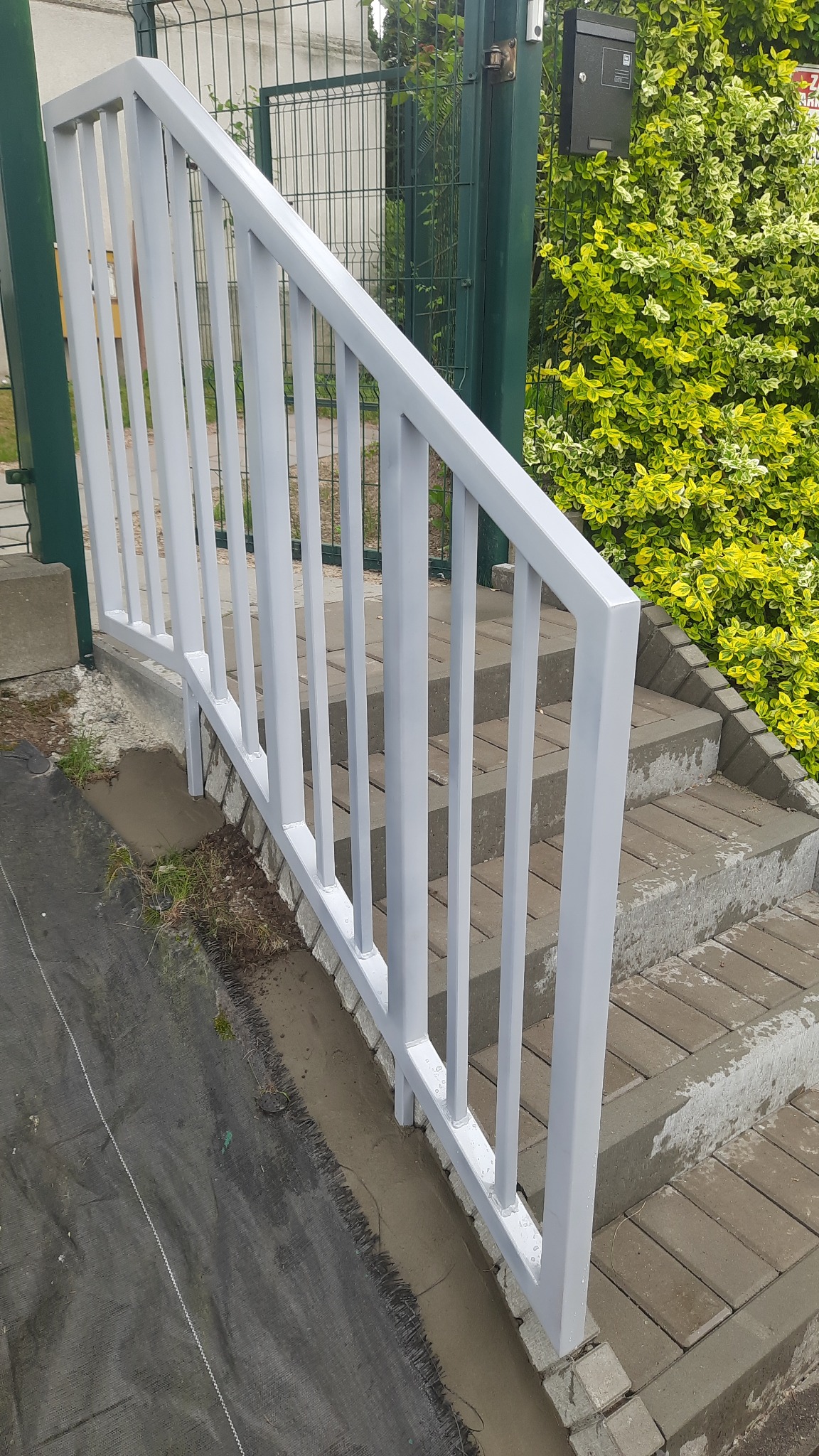 Szara, metalowa balustrada schodowa, widok z dołu, z kostki brukowej, z zielenią w tle. Nowoczesne wykończenie schodów zewnętrznych.