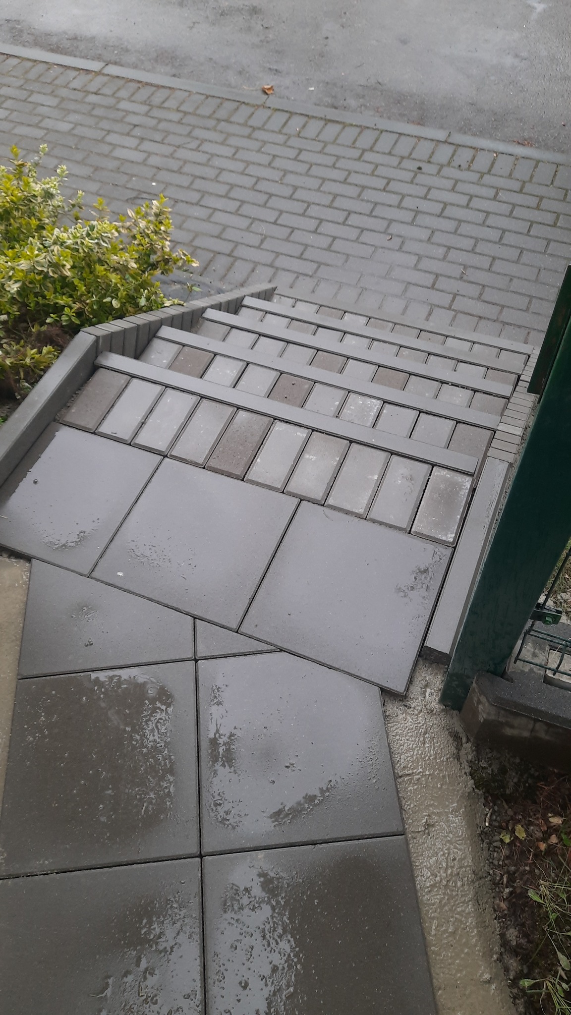 Ułożone schody z kostki brukowej w odcieniach szarości, z widoczną roślinnością obok. Kostka mokra, po deszczu. Widok z góry.