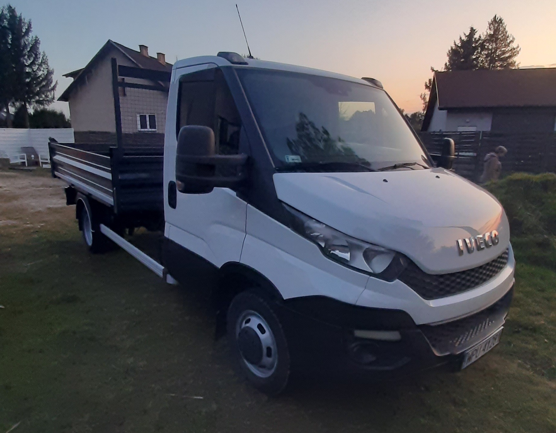 Biały samochód dostawczy Iveco z otwartą skrzynią ładunkową, zaparkowany na trawie, w tle domy i drzewa o zmierzchu, widok z boku.