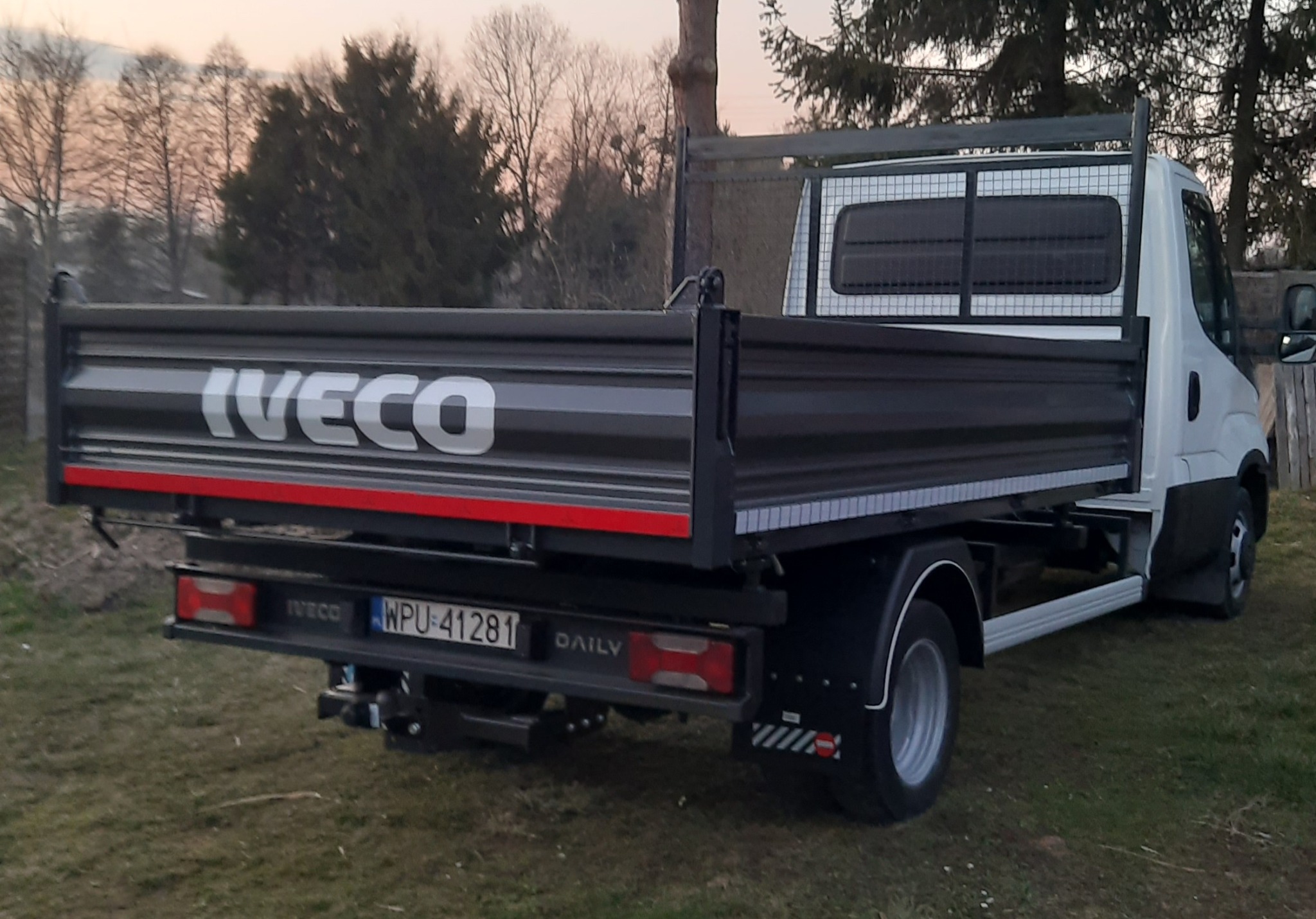 Pojazd Iveco Daily z otwartą skrzynią ładunkową, widok z tyłu na tle zielonej trawy i drzew o zmierzchu. Widoczny numer rejestracyjny i hak holowniczy.