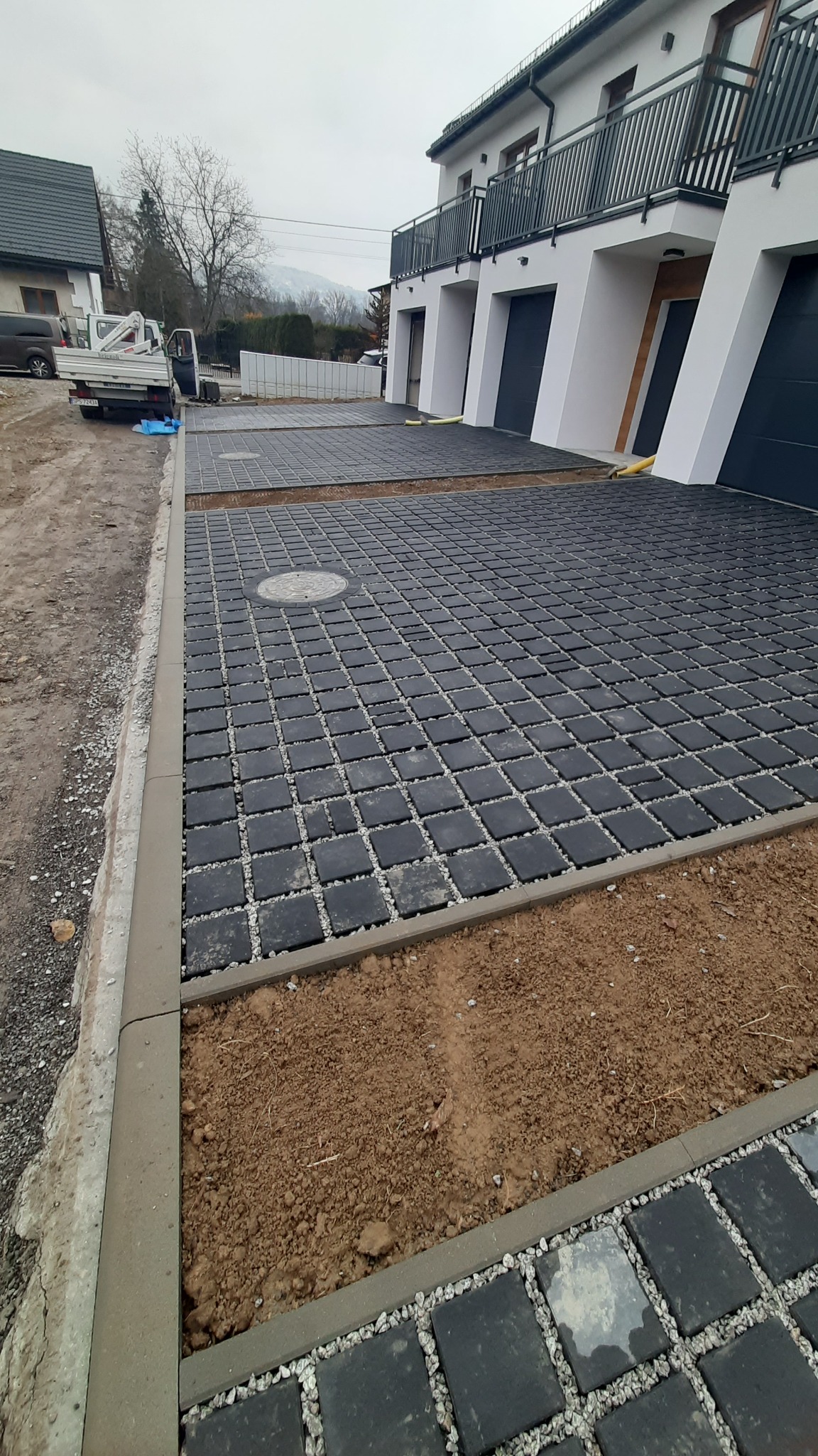Częściowo ułożona, czarna kostka brukowa przed posesją z segmentami. Widoczne obrzeża i przygotowane podłoże. W tle budynek mieszkalny i auto dostawcze.