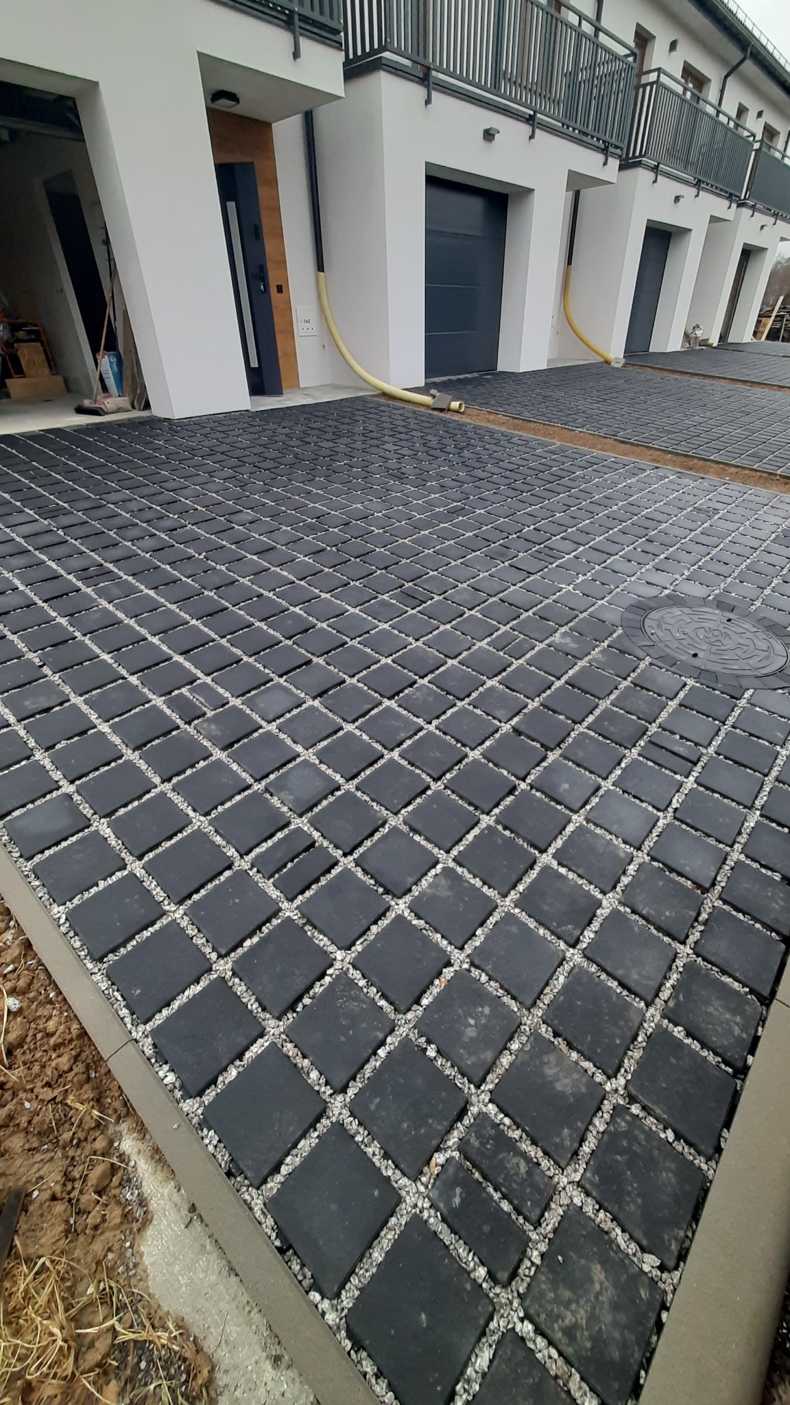 Nowa, czarna kostka brukowa z białym żwirem wypełniającym przestrzenie między elementami, ułożona przed szeregiem nowoczesnych domów.