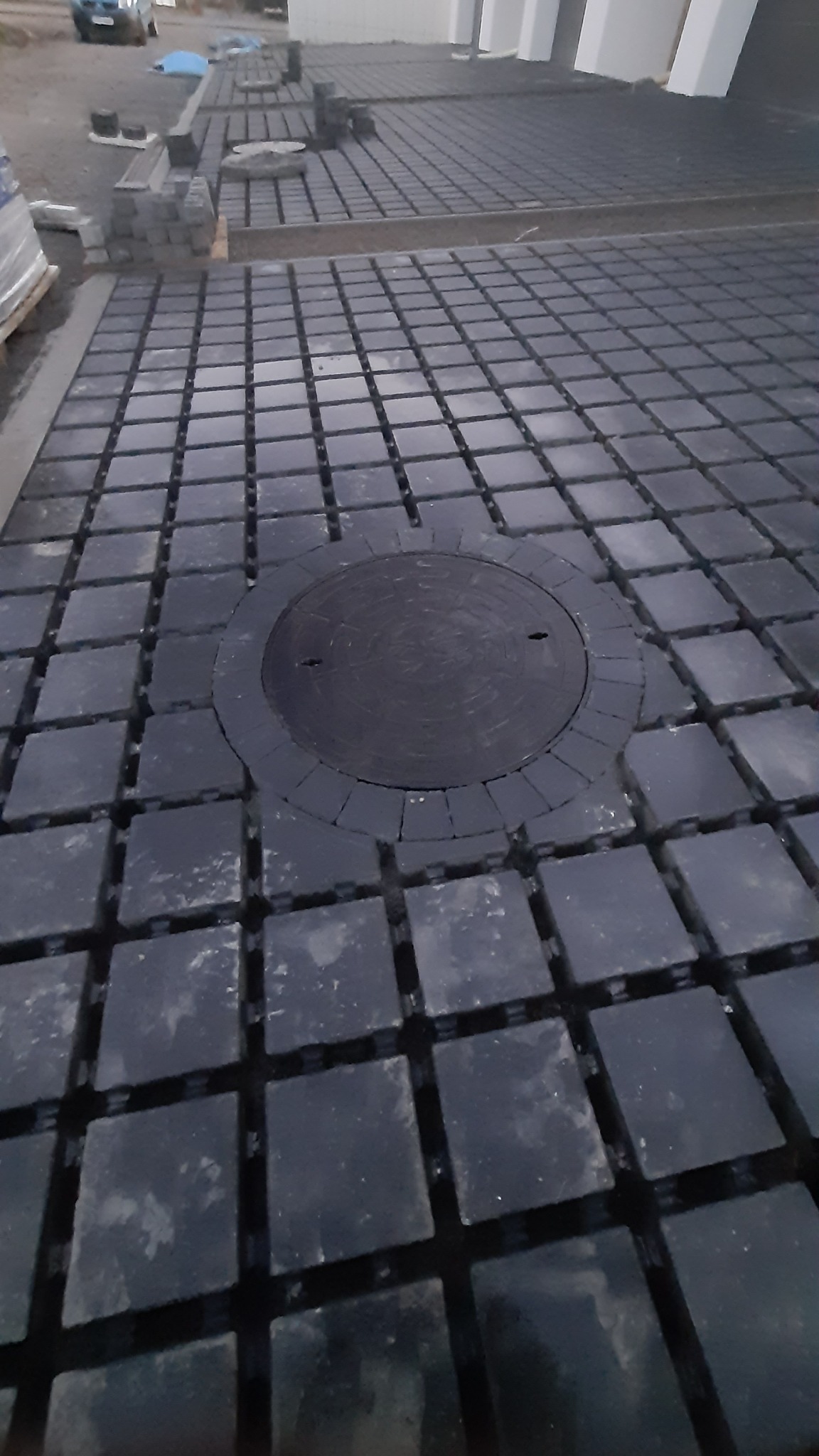 Układanie czarnej kostki brukowej z okrągłą studzienką w centrum. Kostka ułożona na podkładkach dystansowych. Częściowo ułożona kostka w tle.