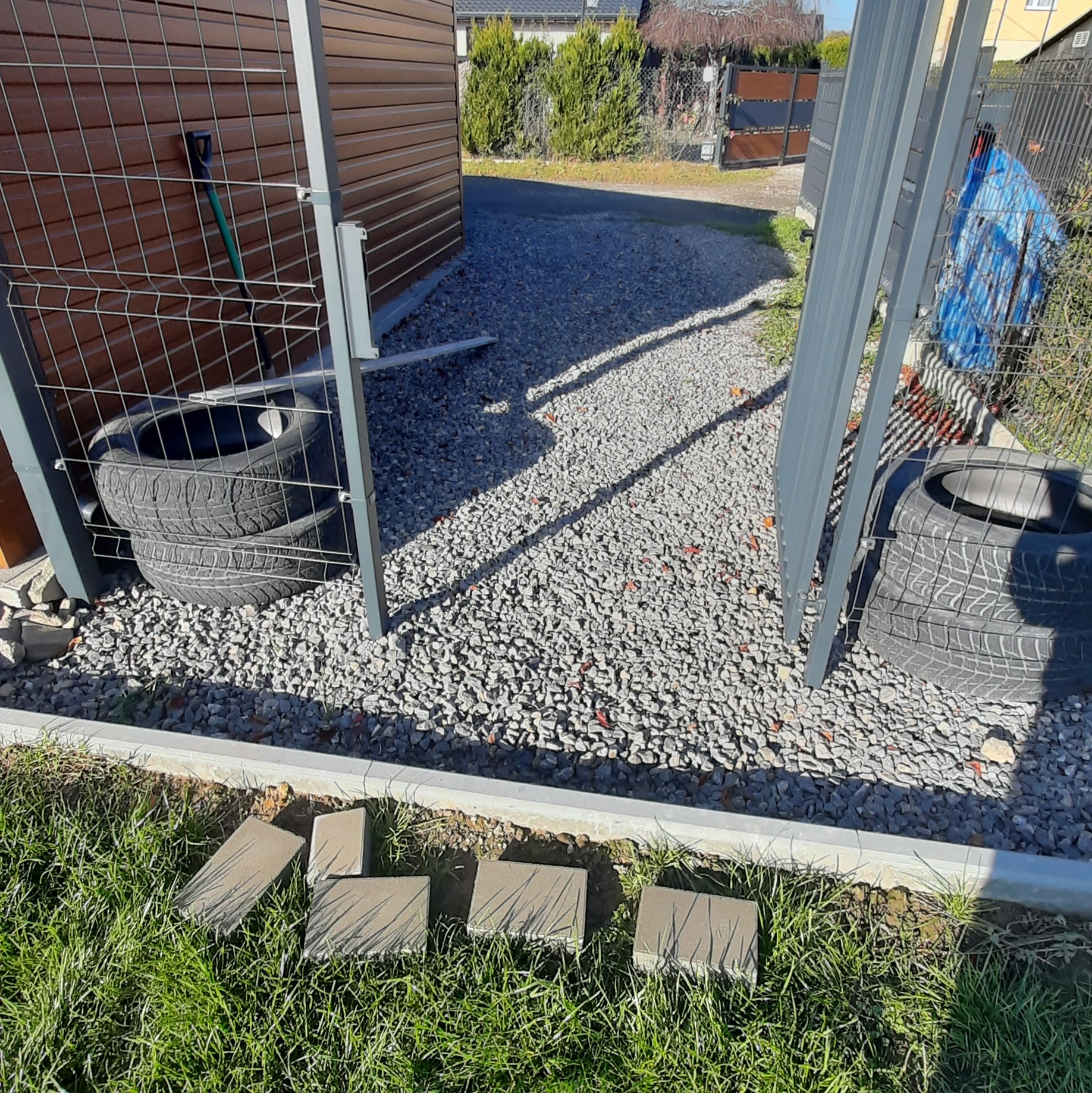 Przygotowanie do układania kostki brukowej: ułożone obrzeża, podsypka z kamienia, kostka leżąca na trawie. Widoczne opony i elementy ogrodzenia.