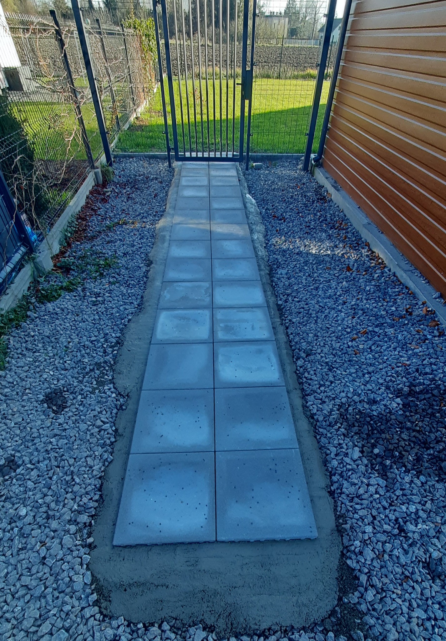 Ścieżka z szarych płyt brukowych prowadząca do furtki, otoczona kamieniami i ogrodzeniem, w tle zieleń trawnika, wykonana wzdłuż brązowej ściany budynku.