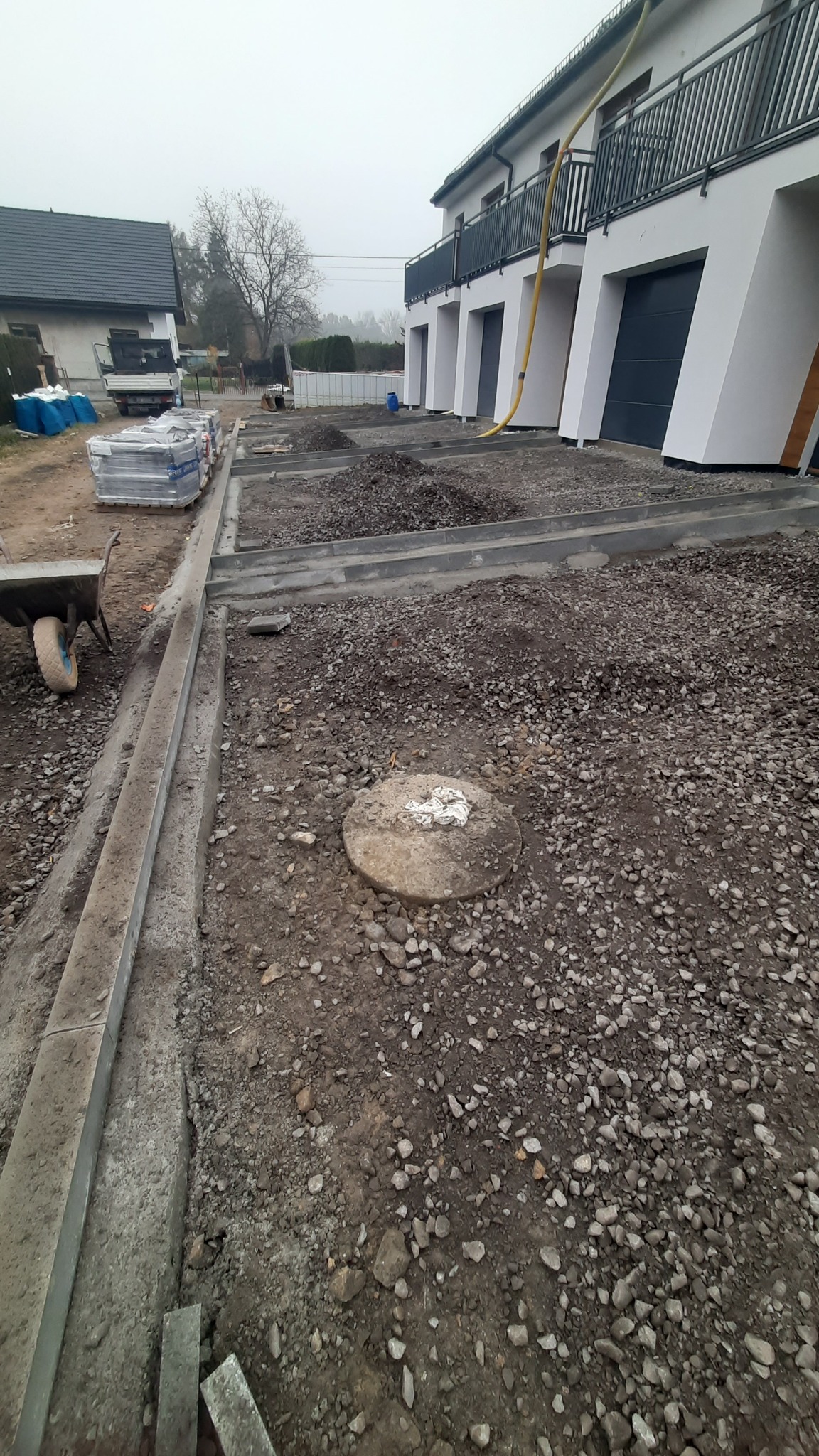 Przygotowanie podłoża pod kostkę brukową: obrzeża ułożone, podsypka rozsypana, widoczna studzienka. Na drugim planie budynek z balkonami i sterta kostki.