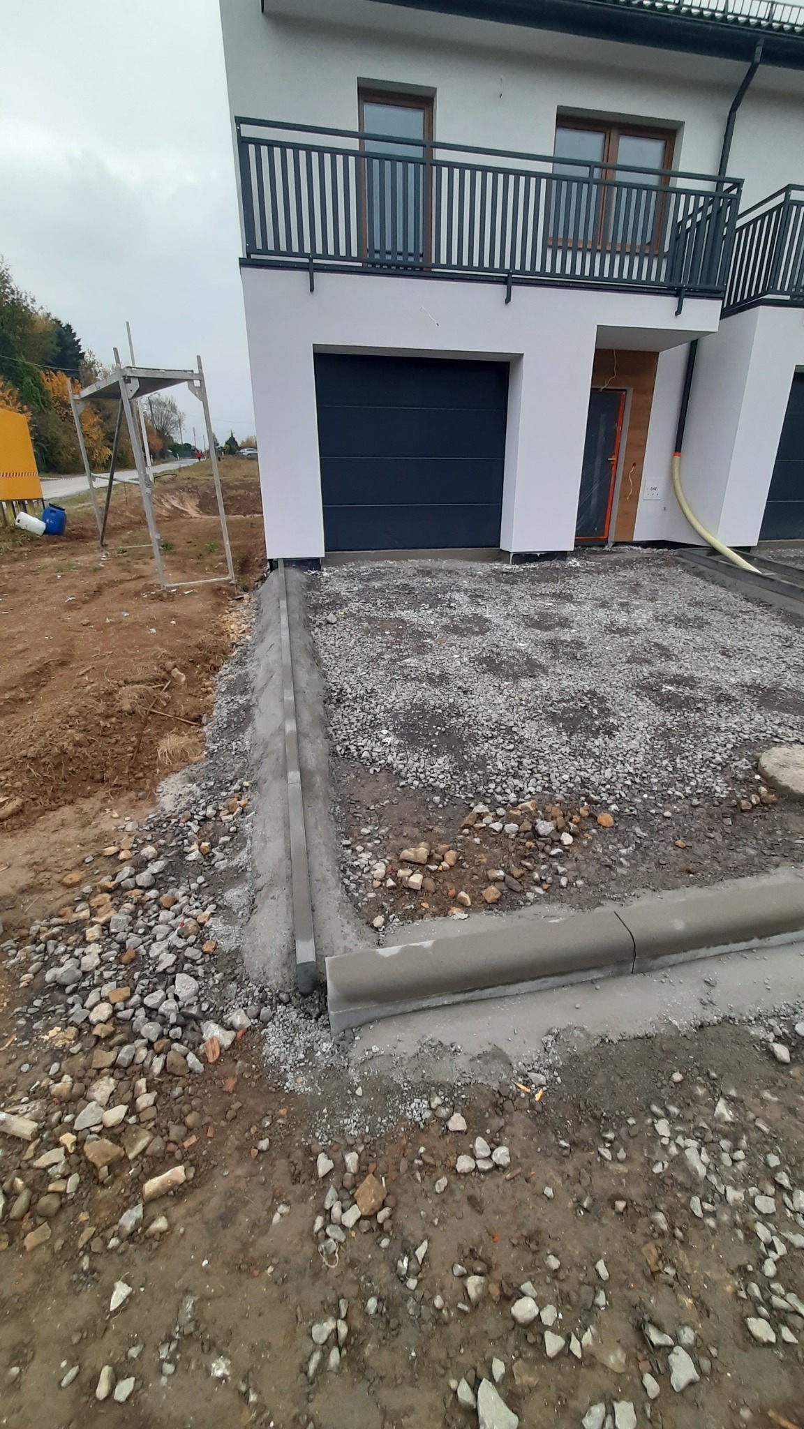 Wyrównany teren pod kostkę brukową przed domem z szarą elewacją i czarnymi bramami garażowymi. Widoczne obrzeża i podsypka z kamienia. Etap przygotowania podłoża.