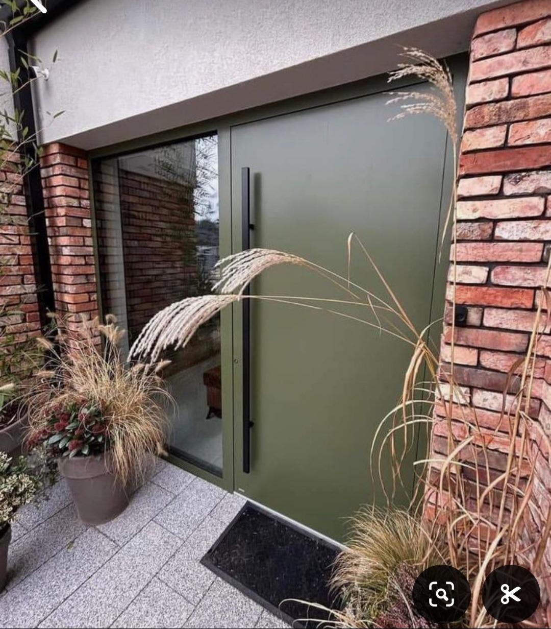 Nowoczesne wejście do domu z oliwkowymi drzwiami, obok ceglana ściana i dekoracyjne trawy. Szara kostka brukowa tworzy eleganckie podejście.