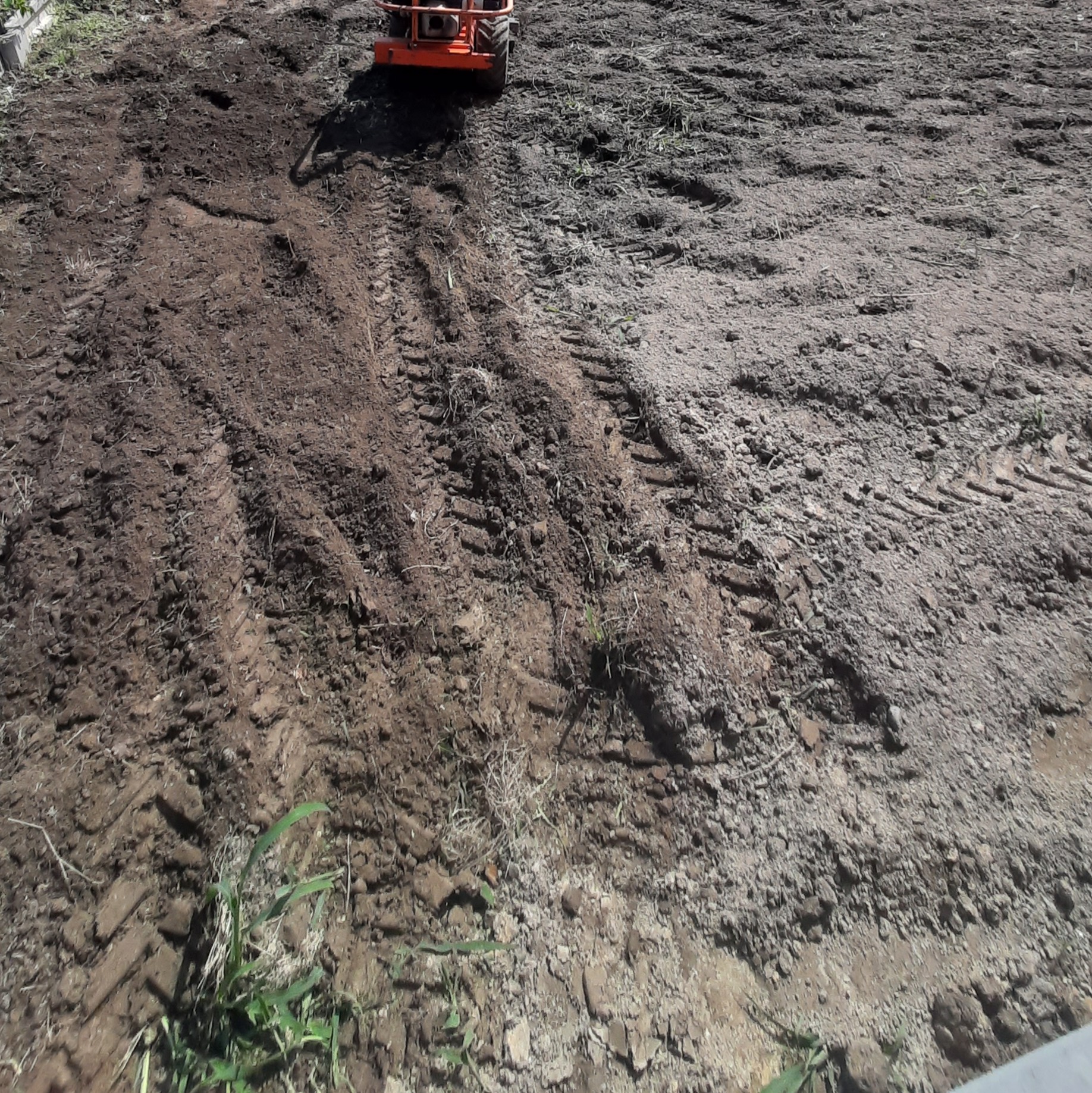 Przygotowanie terenu pod budowę: wyrównana ziemia z widocznymi śladami opon maszyny budowlanej. W tle pomarańczowa maszyna do ubijania gruntu.
