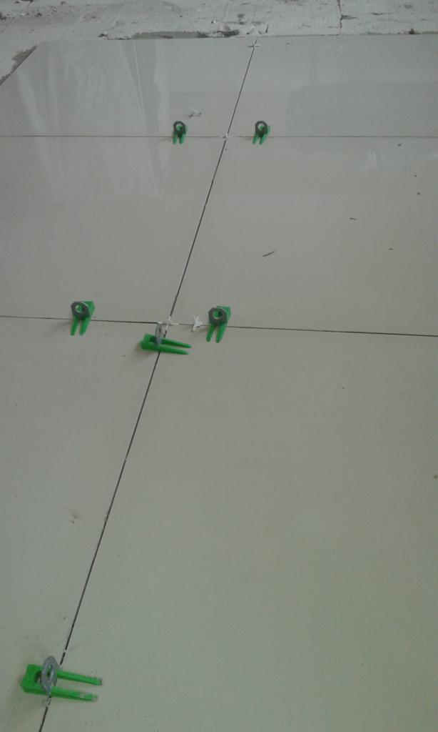 Układanie jasnych, kwadratowych płytek podłogowych z użyciem zielonych klipsów poziomujących, widoczne wąskie fugi.