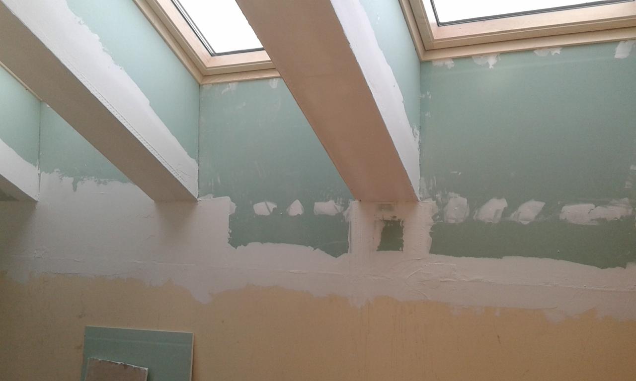 Wnętrze w trakcie prac wykończeniowych: ściany z widocznymi łatami, nałożoną gładzią, oraz drewniane belki pod skosami okien dachowych.