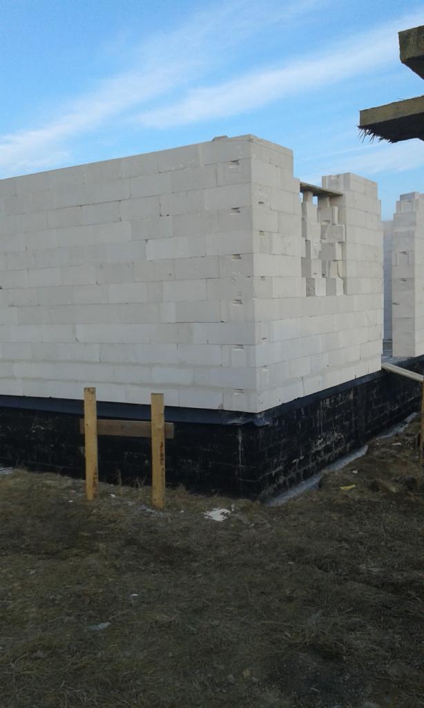 Wznoszenie ściany parteru z białych bloczków na czarnej hydroizolacji fundamentów, widoczne drewniane podpory i fragment nieba.