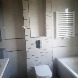 Wykończona łazienka z białymi płytkami imitującymi cegłę, grzejnik drabinkowy i podwieszana toaleta z chromowanym przyciskiem spłukiwania w zabudowie z wnęką.