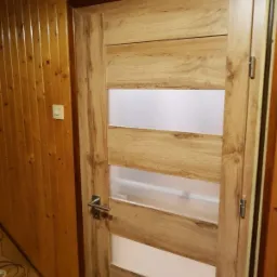 Drewniane drzwi wewnętrzne z matowymi szybami, zamontowane w boazerii, widoczny metalowy kluczyk w zamku.