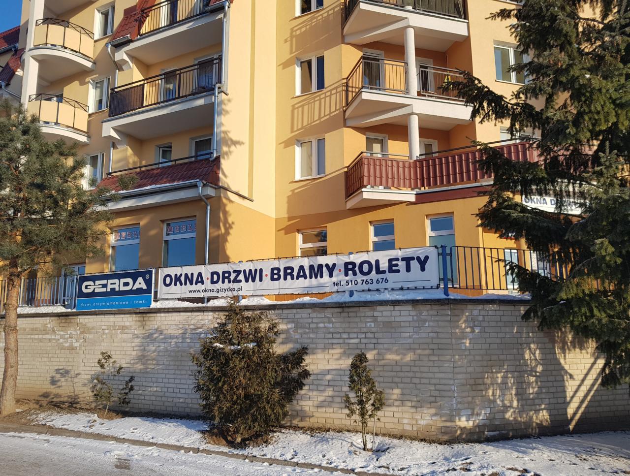 Budynek z żółtą elewacją i balkonami, na murze baner reklamowy firmy Gerda oferującej okna, drzwi, bramy i rolety, widoczne drzewa i śnieg.