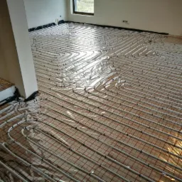 Rozłożona instalacja ogrzewania podłogowego z rurkami na siatce, pokryta przezroczystą folią ochronną, widok z góry w jasnym pomieszczeniu w trakcie montażu.