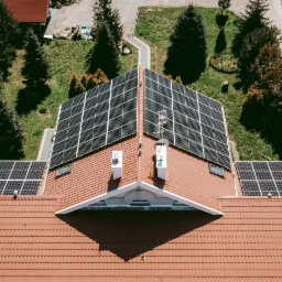 Widok z lotu ptaka na dach domu z czerwoną dachówką, na którym zamontowane są panele słoneczne; widoczne kominy, drzewa i fragment pola w tle.