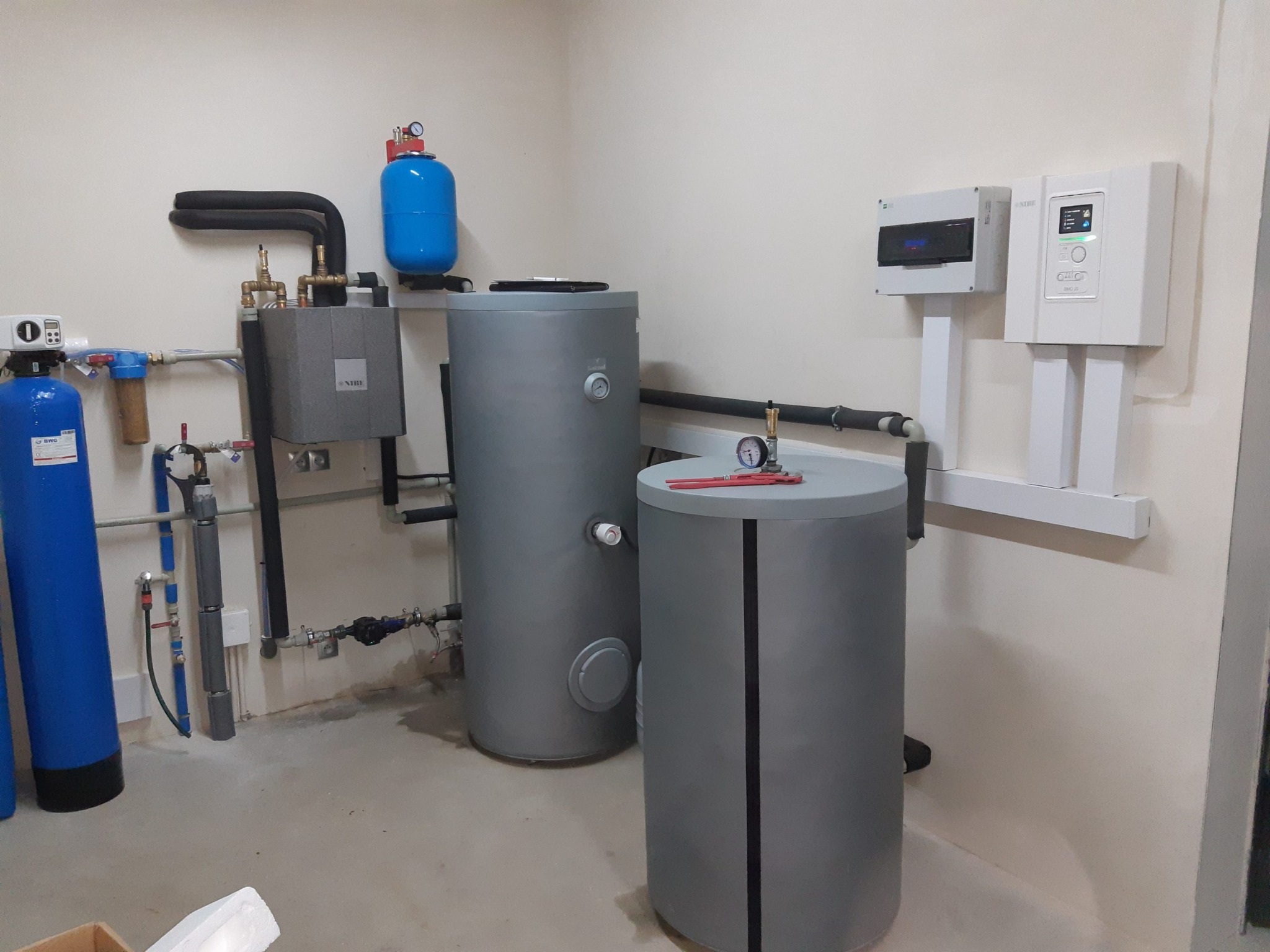 Kompletna instalacja grzewcza z dwoma szarymi zbiornikami, niebieskim zbiornikiem wyrównawczym, filtrami wody i panelem sterowania na ścianie, widoczne rury i zawory, na górze zbiornika leżą...