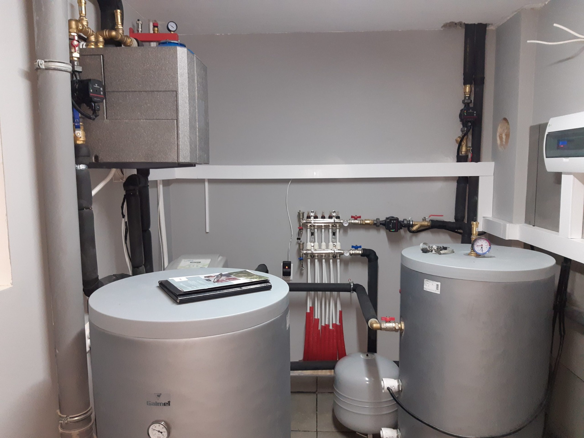 Węzeł cieplny z dwoma zbiornikami Galmet, rozdzielaczem, pompą obiegową i automatyką, widoczne rury instalacyjne i manometry, na jednym zbiorniku leżą prospekty reklamowe.