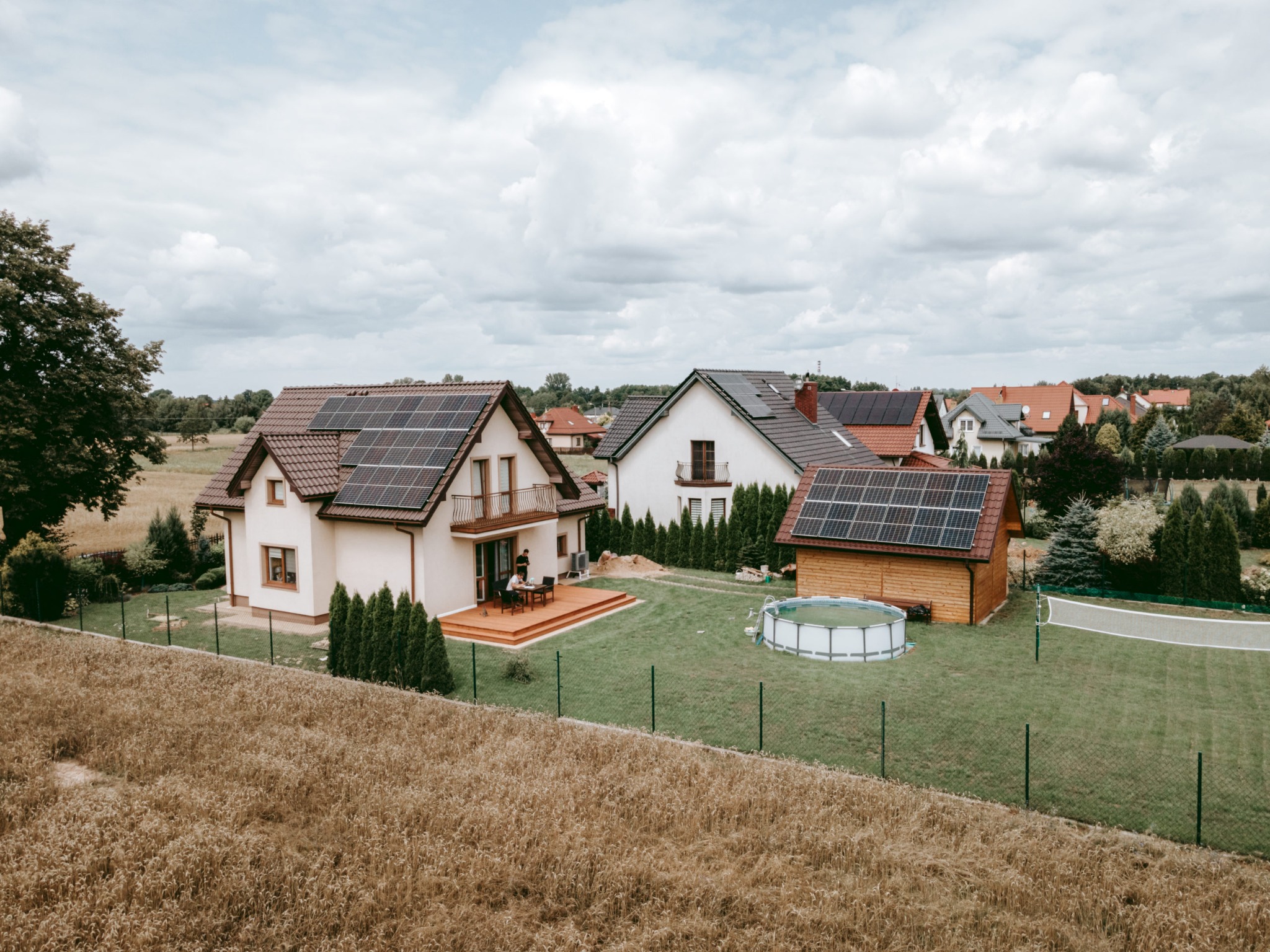 Widok z góry na domy jednorodzinne z panelami słonecznymi na dachach w słoneczny dzień, na trawniku basen oraz siatka do gry w siatkówkę.