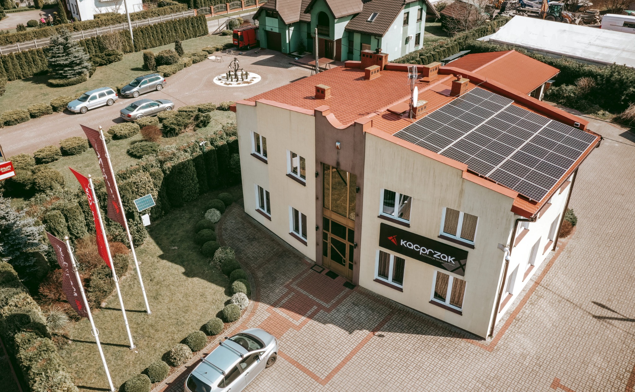 Widok z góry na budynek z panelami słonecznymi na dachu, z logo firmy 'Kacprzak' na elewacji, otoczony zadbanym ogrodem i brukowanym podjazdem, na którym stoi srebrny samochód.