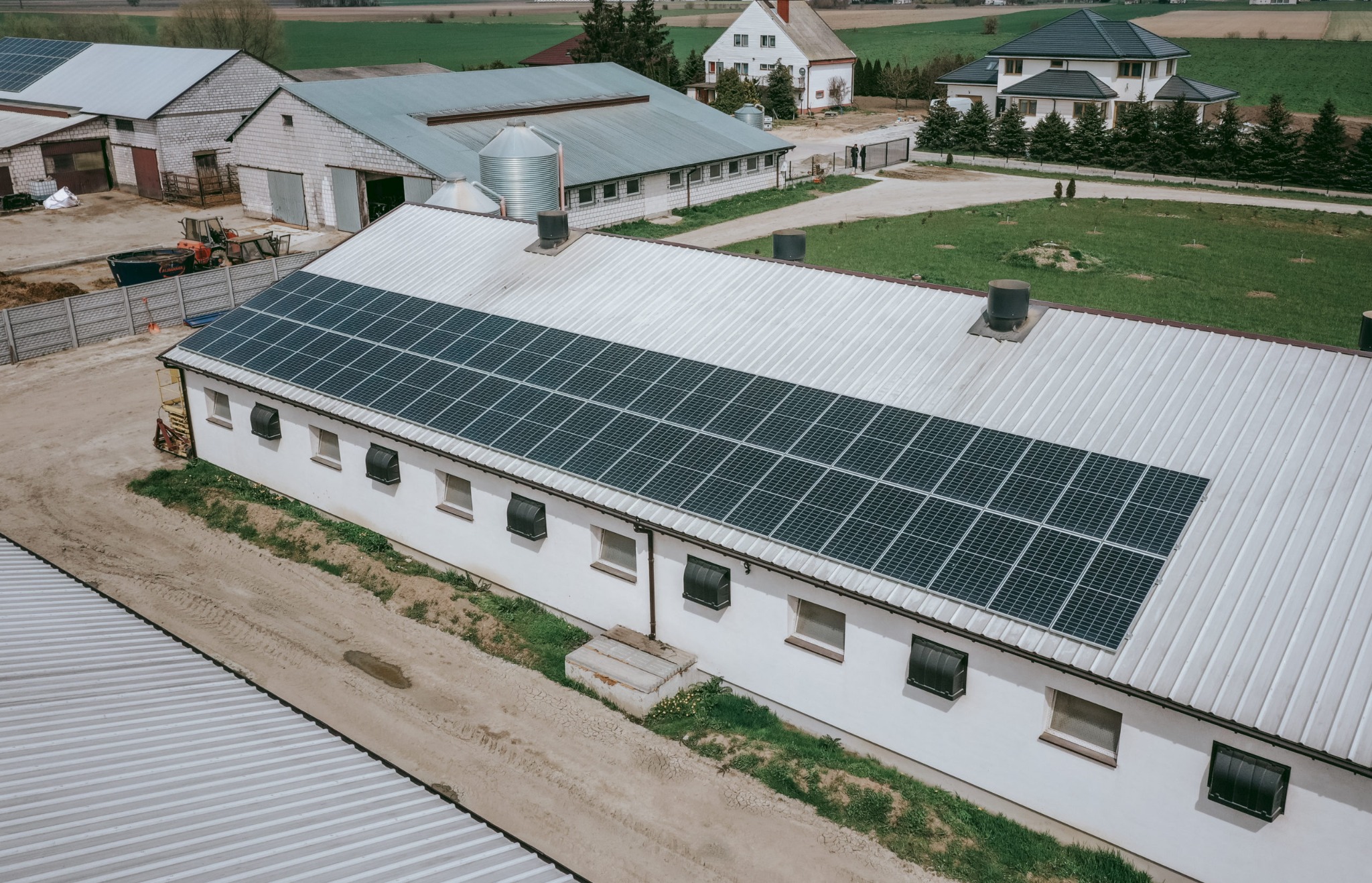 Widok z góry na instalację paneli słonecznych na dachu budynku gospodarczego na terenie wiejskim, z widocznymi oknami i elementami wentylacyjnymi na dachu, w tle zabudowania gospodarcze i domy.