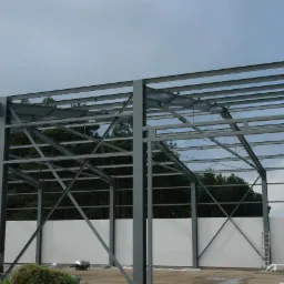 Szkielet konstrukcji stalowej hali w trakcie budowy, widoczne elementy montażowe, białe płyty ścienne, las w tle i pomarańczowy samochód dostawczy w oddali.