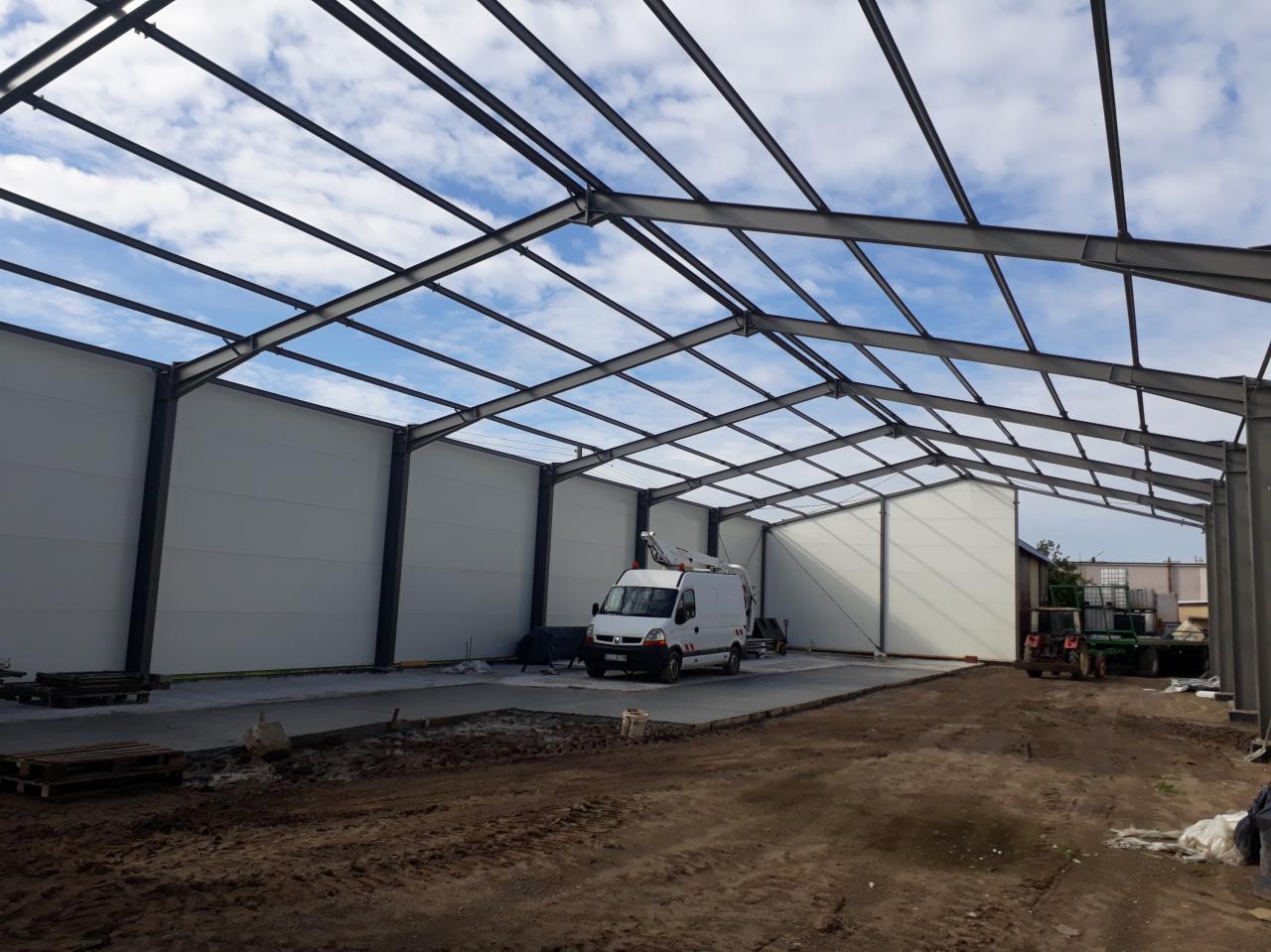 Wnętrze hali w trakcie budowy z widoczną stalową konstrukcją dachu i ścianami z paneli warstwowych, z zaparkowanym białym samochodem dostawczym z podnośnikiem na zewnątrz i przygotowanym podłożem...